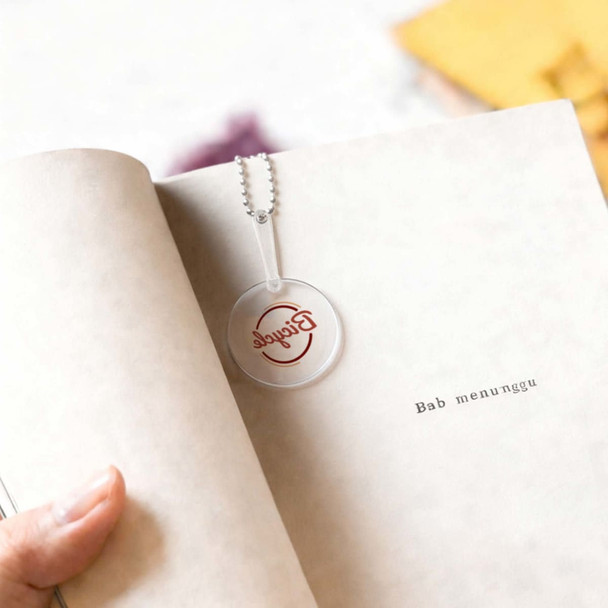 A transparent PVC hand fan with a keychain, featuring a circular shape and a branding logo, rests on a book page.