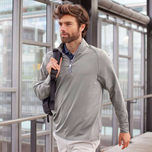 A man wearing a light grey half-zip sweatshirt, carrying a shoulder bag, walking in a modern building.