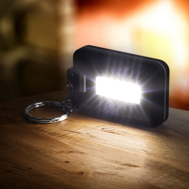 A compact black key ring light emits a bright white light, resting on a wooden surface with a blurred background.