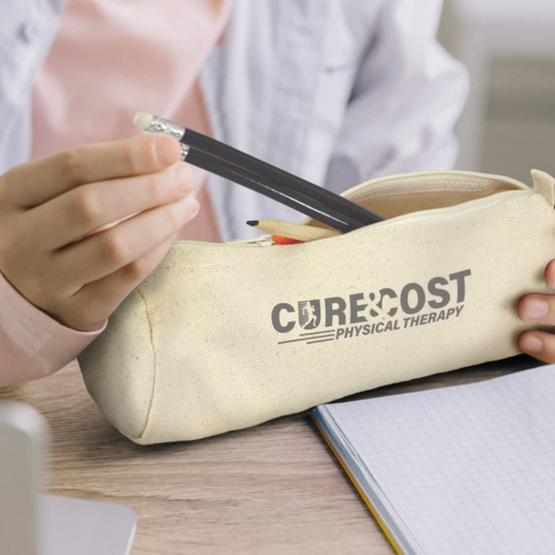 A cream pencil case with a logo, containing pencils and a pen, placed on a wooden surface next to a notebook.