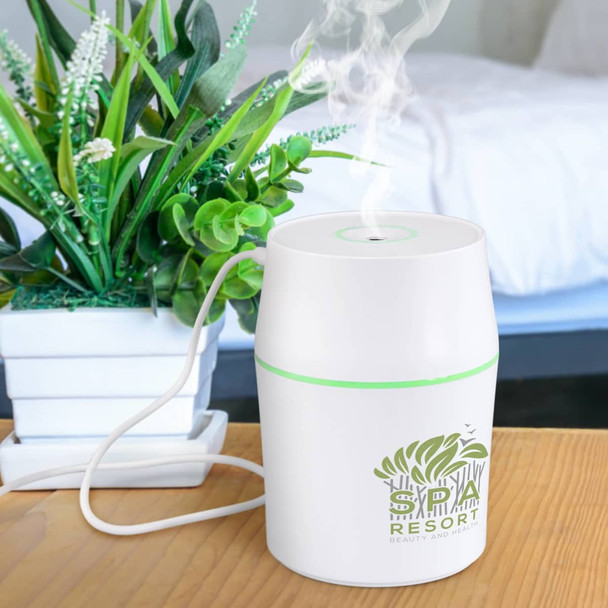 A white aroma diffuser with a green accent, emitting steam, placed on a wooden table near a potted plant.