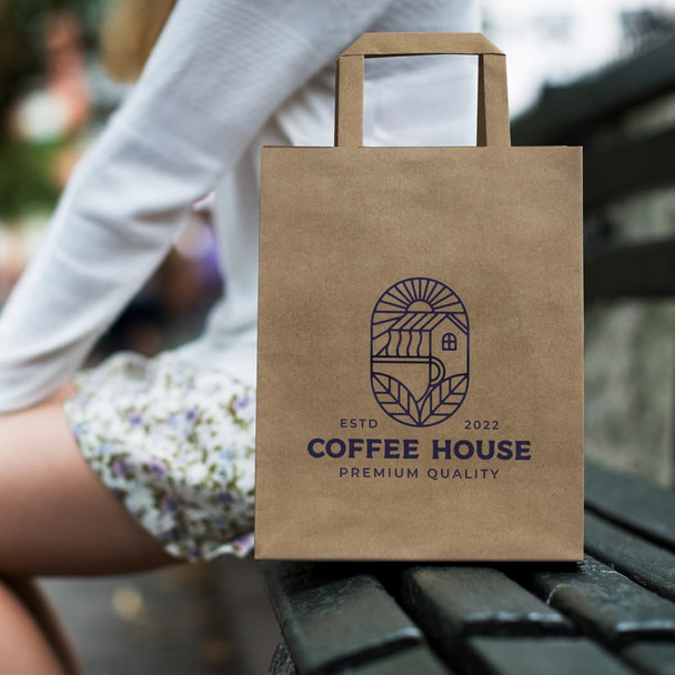 A medium flat handle paper bag in brown with a logo, placed on a bench beside a person.