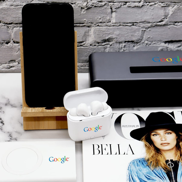 A bamboo phone stand displays a phone, alongside white earbuds in a case and a magazine on a marble surface.