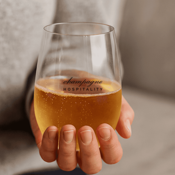 A stemless shatterproof red wine glass filled with sparkling golden beverage, held in a person's hand. Features a logo.