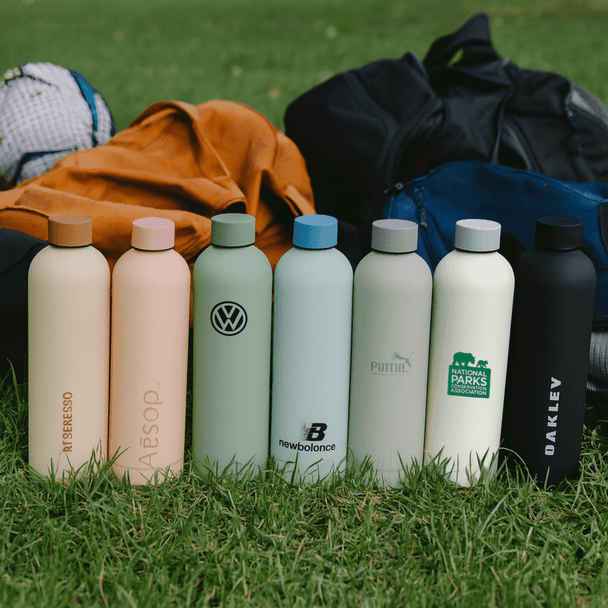 A lineup of drink bottles in pastel shades, featuring a variety of designs and logos, set on grass with bags in the background.