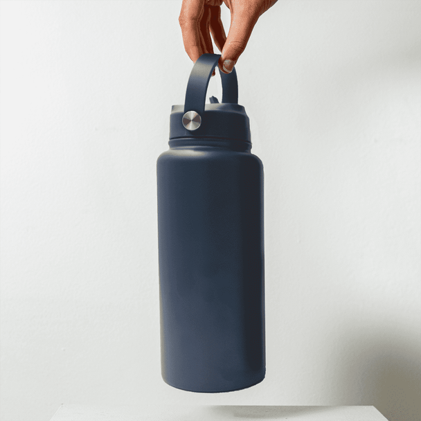 A drink bottle in dark blue, featuring a handle and a silver accent, held by a hand against a plain background.
