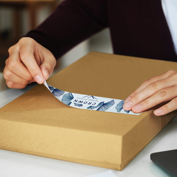A person applying a blue and white sticker to a brown packaging box. The box is on a flat surface.