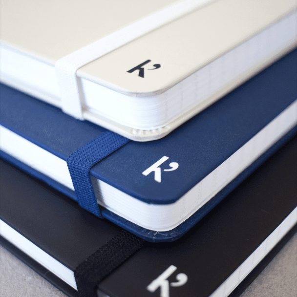A stack of A5 hardcover notebooks in white, navy blue, and black, featuring an elastic band and a logo.