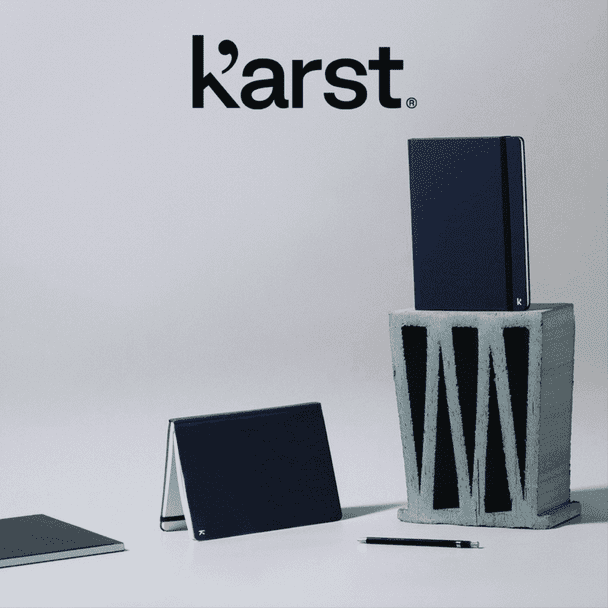 A navy hardcover notebook stands on top of a grey block, accompanied by a smaller notebook and a pen. It has a logo.