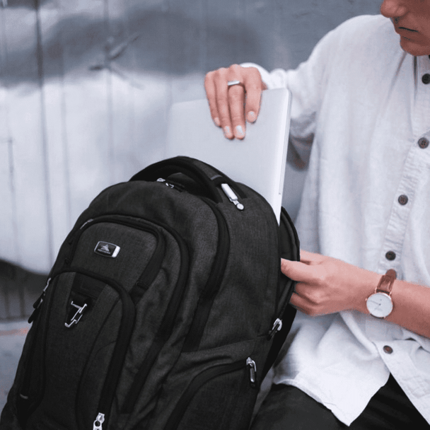 A dark grey laptop backpack with multiple compartments, sitting beside a person holding a laptop. The backpack features a logo.