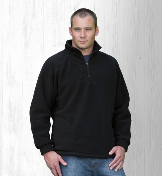 A man wearing a black polar fleece pullover with a half zip, standing against a light wooden background.