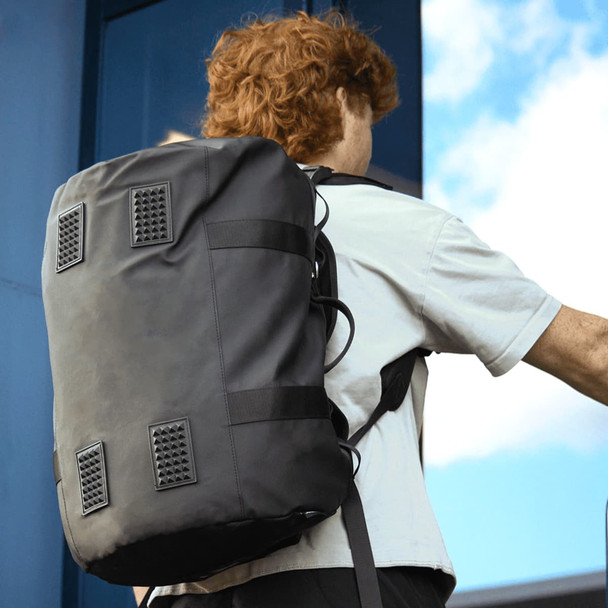 A black duffel bag with a textured base and adjustable straps, worn on a person's back against a blue background.