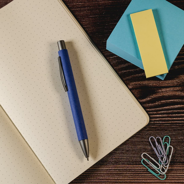A blue pen placed on a dotted notebook, accompanied by sticky notes and paperclips. A blue pen placed on a dotted notebook, accompanied by sticky notes and paperclips.