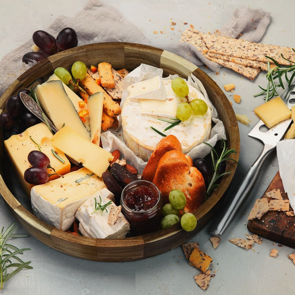 A wooden platter tray filled with various cheeses, grapes, crackers, and a jar of jam, styled with herbs.