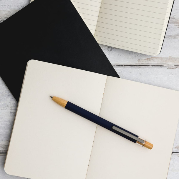 A metal pen with a blue barrel and wooden accents resting on an open notebook with blank pages. A metal pen with a blue barrel and wooden accents resting on an open notebook with blank pages.