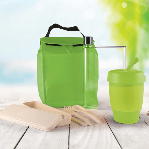 A green picnic bag contains a drink bottle, reusable cup, utensils, and food containers, set against a blurred outdoor background.