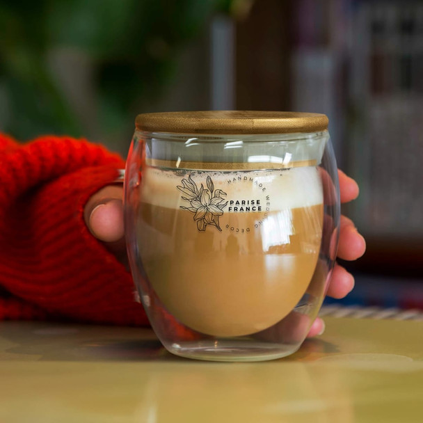 A glass cup with a wooden lid, featuring a frothy coffee inside, held in a hand with a red sweater.