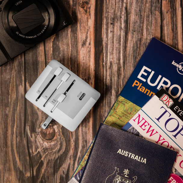 A white travel adaptor positioned on a wooden surface, surrounded by a camera, travel books, and a passport.