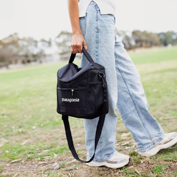 A black cooler bag with a strap, held by a person in light blue jeans, set against a grassy outdoor background.