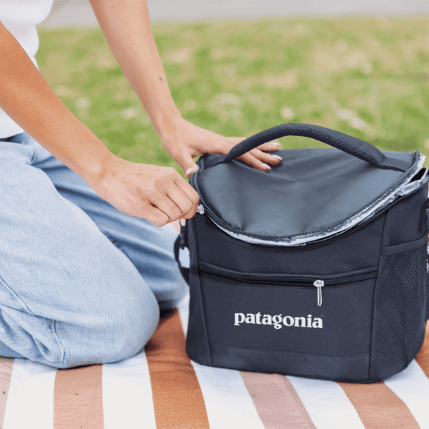 A black cooler bag with a zippered top, featuring a handle and a logo, placed on a picnic blanket.