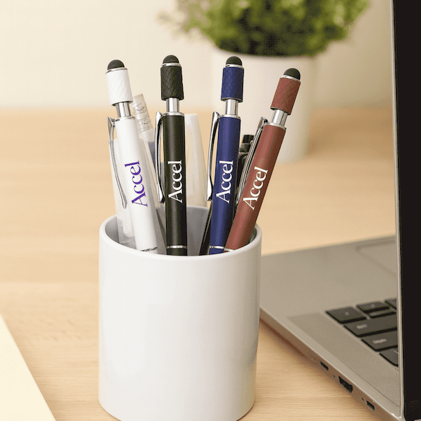 A pen holder featuring several metal pens in white, black, blue, and maroon, placed near a laptop and plant.