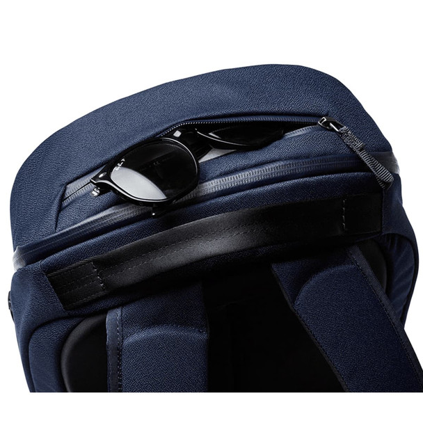 A navy Bellroy Via Backpack with a zippered compartment featuring sunglasses on top. The bag has a sleek, textured finish.
