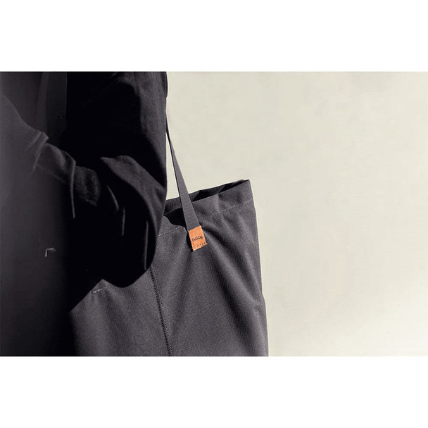 A dark grey tote bag with a branding tag, held by a person in black clothing against a neutral background.