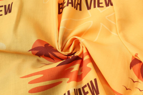 A close-up of a yellow fabric with a beach view design featuring palm trees and waves. It has a logo in the pattern.