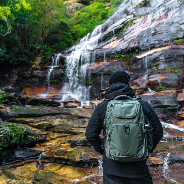 A person stands by a waterfall, wearing a green High Sierra Access 4.0 backpack. The surroundings are lush and rocky.