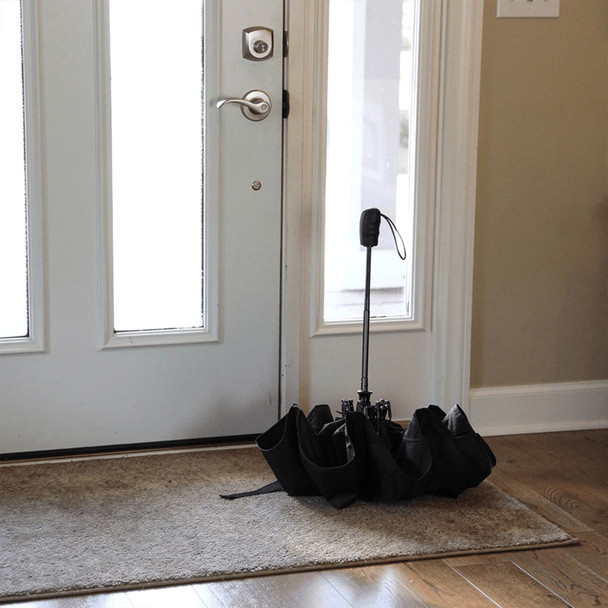 A black recycled tipless umbrella standing upright by a door on a mat. The umbrella has a sleek design and features a logo.