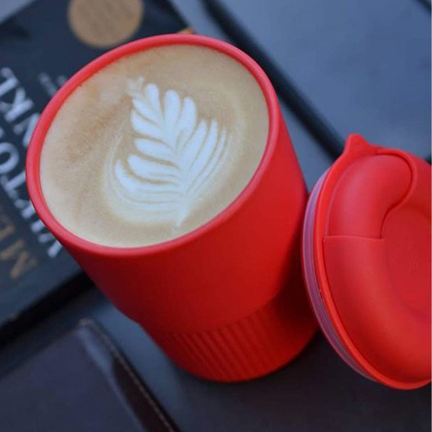 A red reusable coffee cup with a latte featuring leaf art, alongside a dark background with a book.