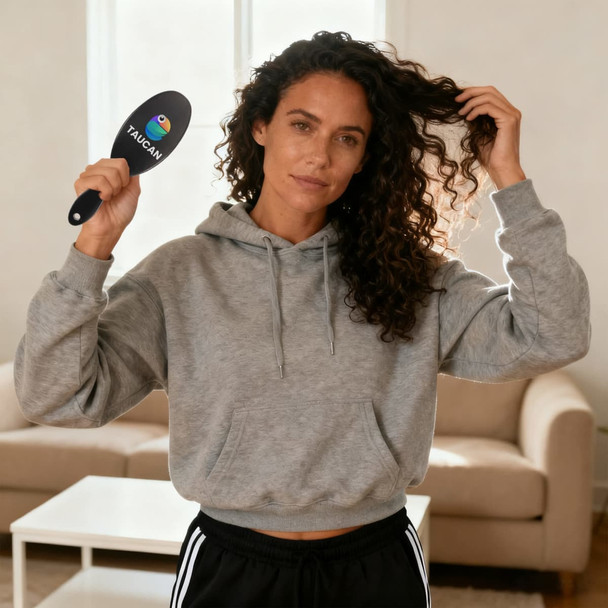 A woman with curly hair holds a black hair brush with a logo, wearing a grey hoodie in a bright room.