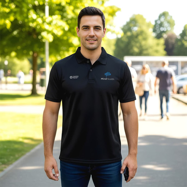 A man wearing a black polo shirt with a logo stands outdoors on a pathway lined with trees.