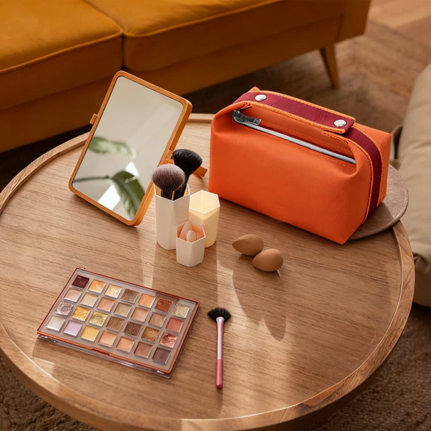 An orange portable canvas cosmetic bag sits on a wooden table with makeup brushes, sponges, a mirror, and an eyeshadow palette.