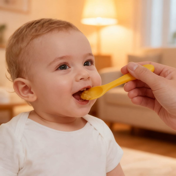 A hand feeds a baby with a yellow silicone spoon in a cosy indoor setting. Soft lighting creates a warm atmosphere.
