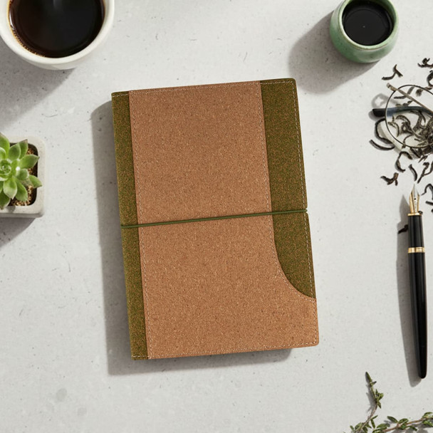 A cork notebook in brown and green, placed on a light surface with a cup, plant, and pen nearby.