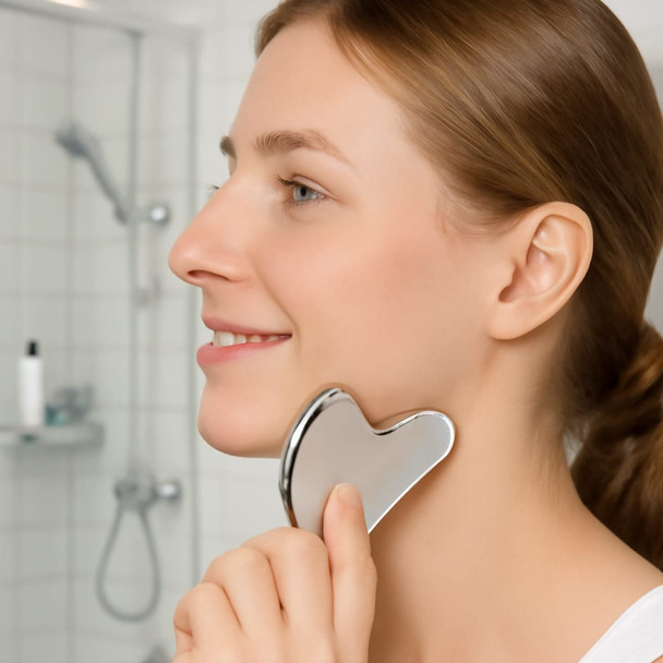A stainless steel Gua Sha massage tool being used on a woman's jawline, featuring a shiny silver finish.
