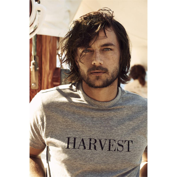 A man wearing a grey crew neck T-shirt featuring a logo. He has long, tousled hair and a beard.