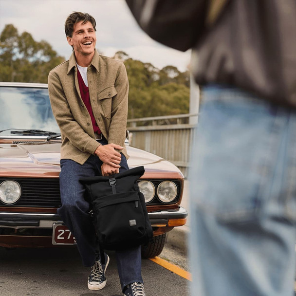 A man in a beige jacket and red sweater sits on a vintage car, holding a black Bellroy Classic Rolltop backpack.