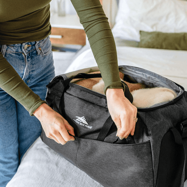 A person opens a charcoal grey duffel bag, revealing a cozy interior with a blanket inside. The bag has a logo.
