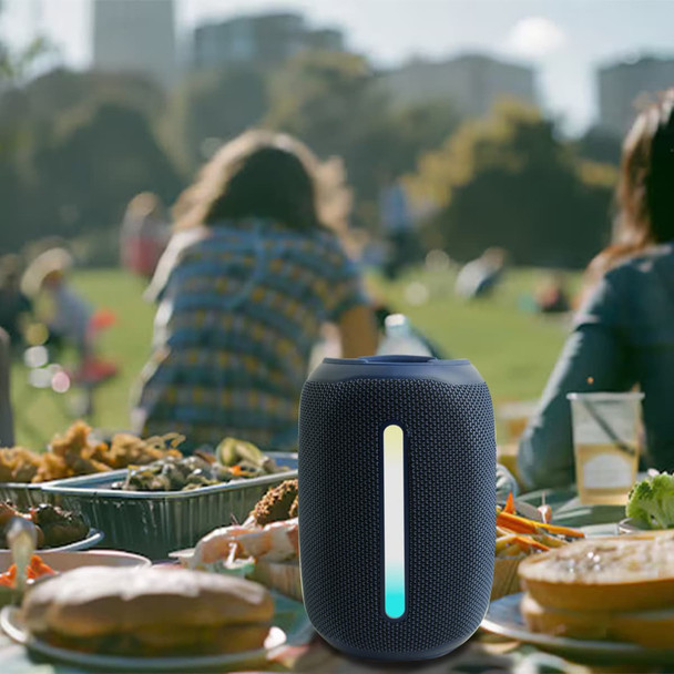 A black outdoor Bluetooth speaker with an LED light strip, set on a picnic table amidst food and drinks.