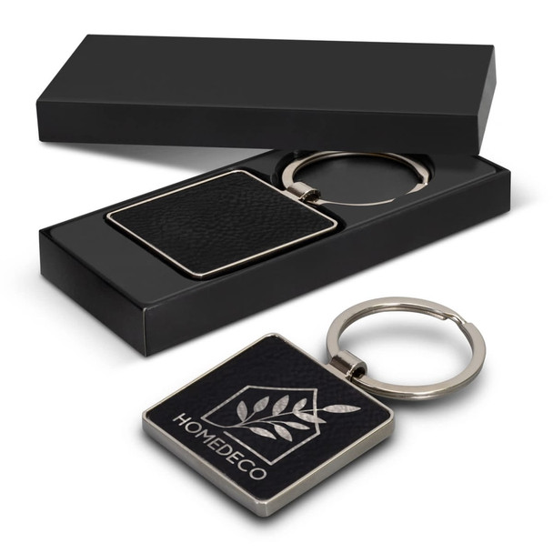A square key ring with a leather-like texture, presented in a black box with a logo.