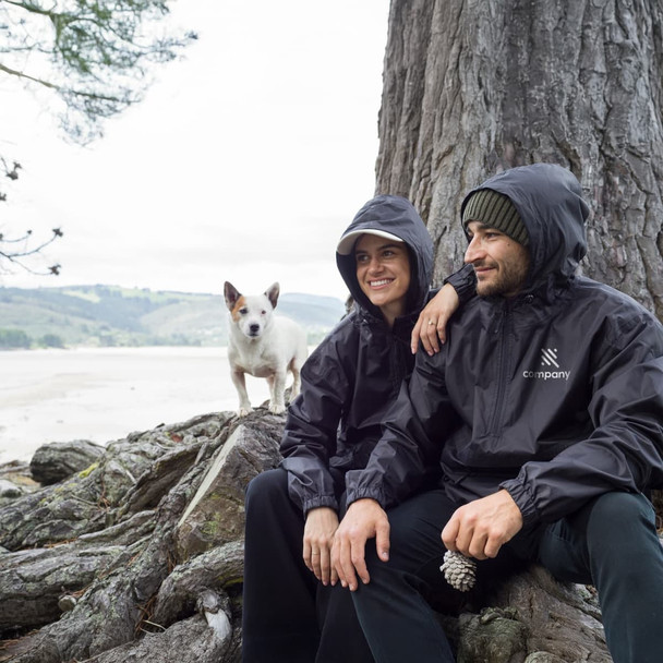 A black windbreaker worn by two people sitting by a tree, with a small dog nearby. The jackets have a logo on them.