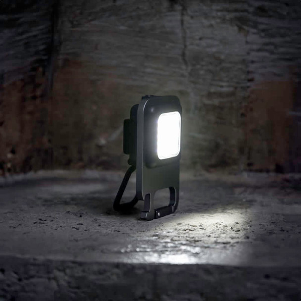 A compact black multi-tool LED light positioned on a textured surface, with a bright light emanating from its front.