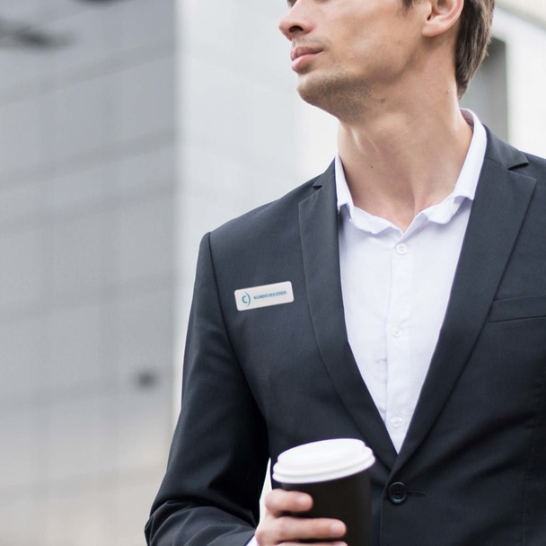 A man in a black suit holding a coffee cup, wearing a custom acrylic name badge on his lapel.