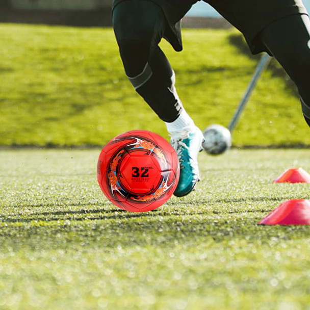 A vibrant red soccer ball with black and orange designs on grass, with a person in action nearby.
