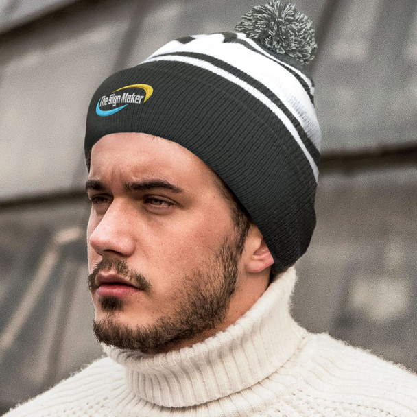 A black and white beanie with a pom pom, worn by a young man in a neutral sweater, features a logo.