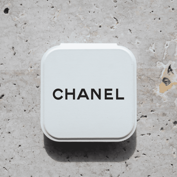 A compact mirror in a white case with a logo on the top, set against a textured stone background.