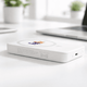 A white wireless charging power bank with a logo, resting on a desk near plants and a laptop.