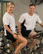 A white and navy short sleeve contrast tee worn by a man and woman sitting on stationary bikes in a gym.